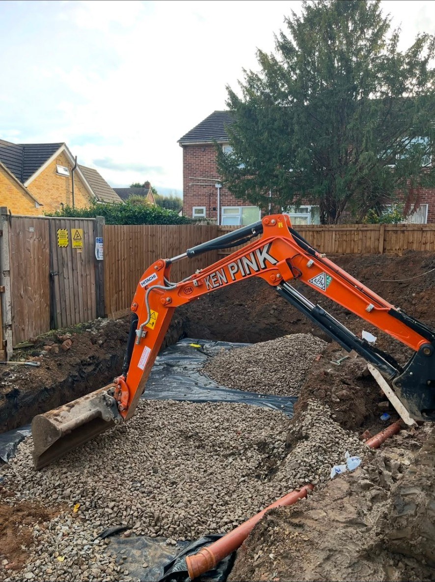 Orange digger arm over garden excavation site with sub-base stone and drainage pipes