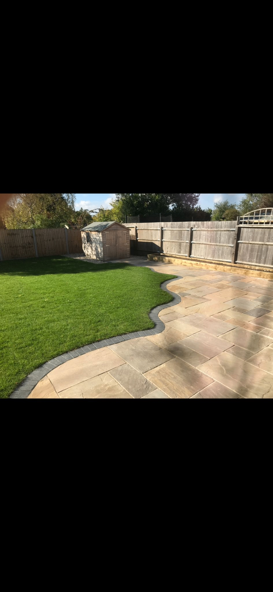 Sweeping autumn-blend Indian sandstone patio with charcoal block edge curving around a freshly laid lawn.