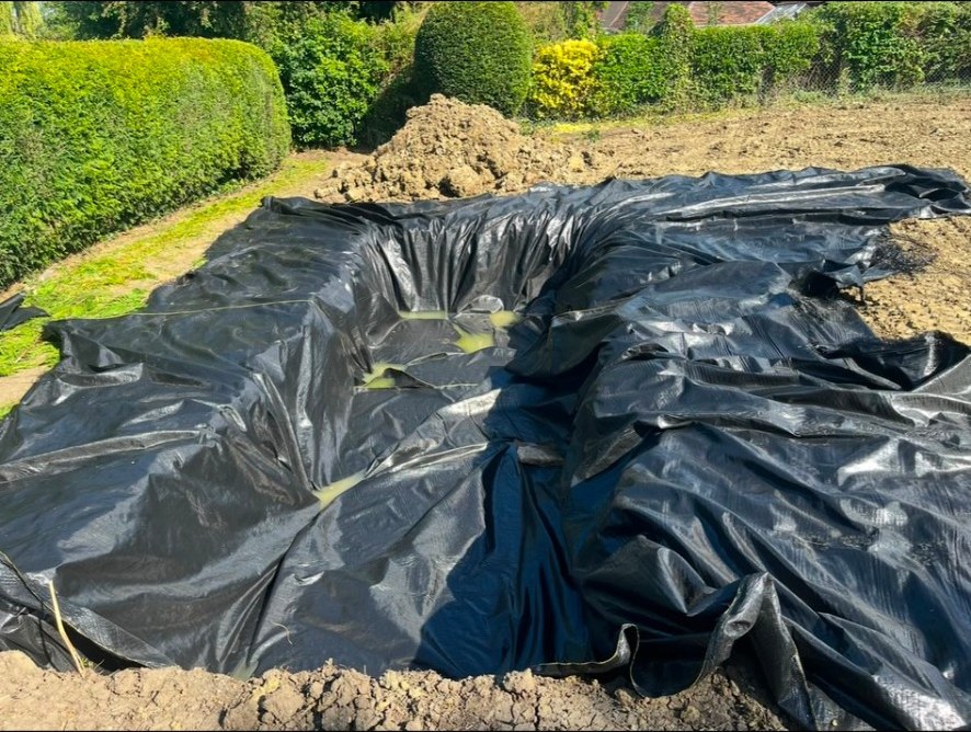 Excavated ground with black pond liner installed and soil mounds in a garden.