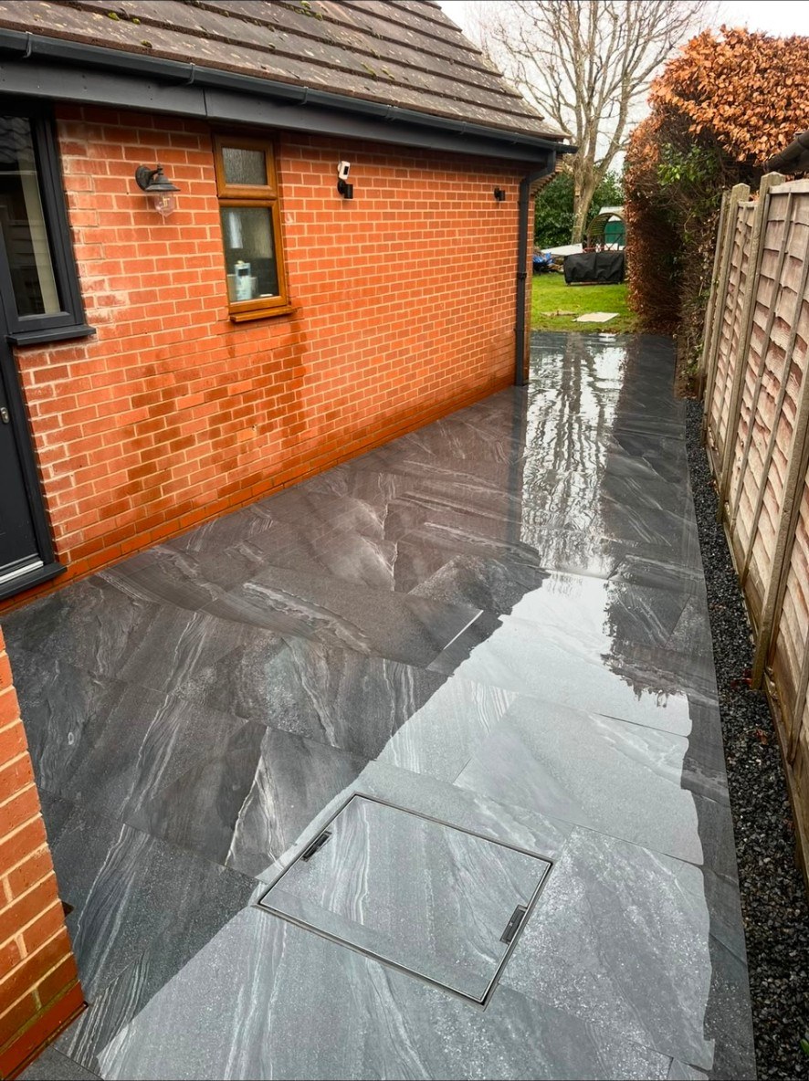 Dark grey porcelain tiled side path with matching recessed manhole cover against red brick house.