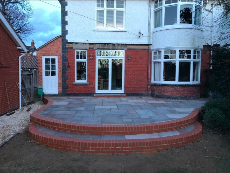 Sandstone patio with curved red brick steps and border in a residential back garden.