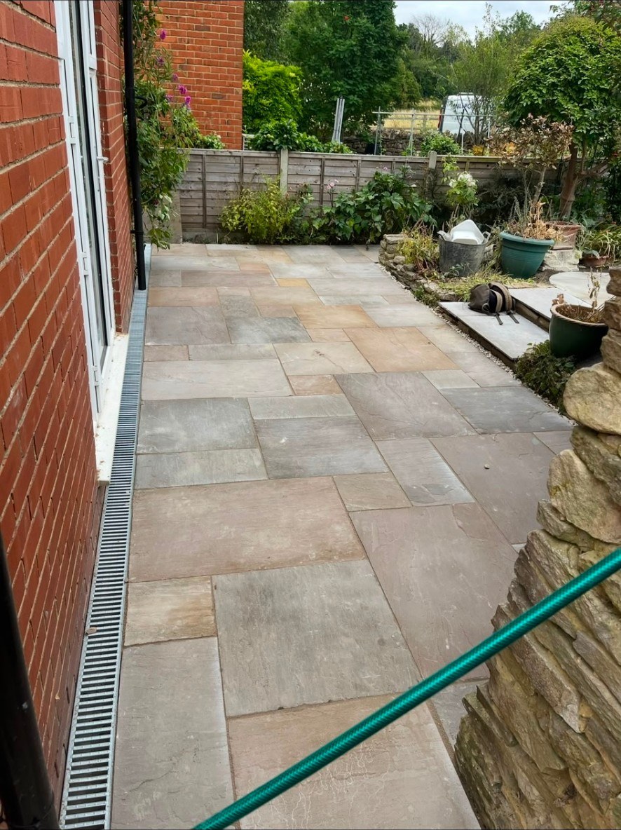 Natural riven sandstone patio with drainage channel besides a red brick house.