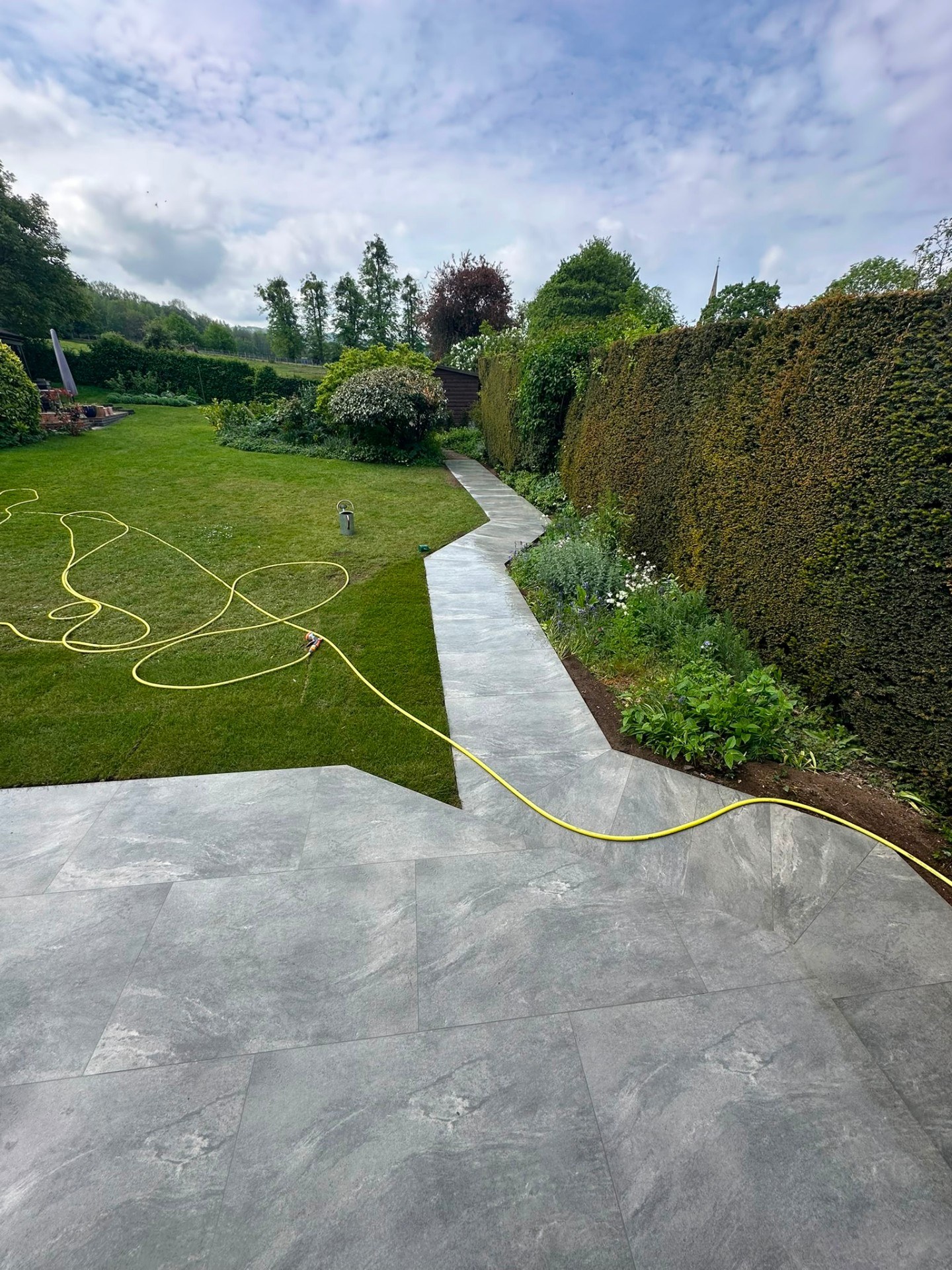 Modern grey porcelain patio and garden path alongside a lawn and tall hedge.