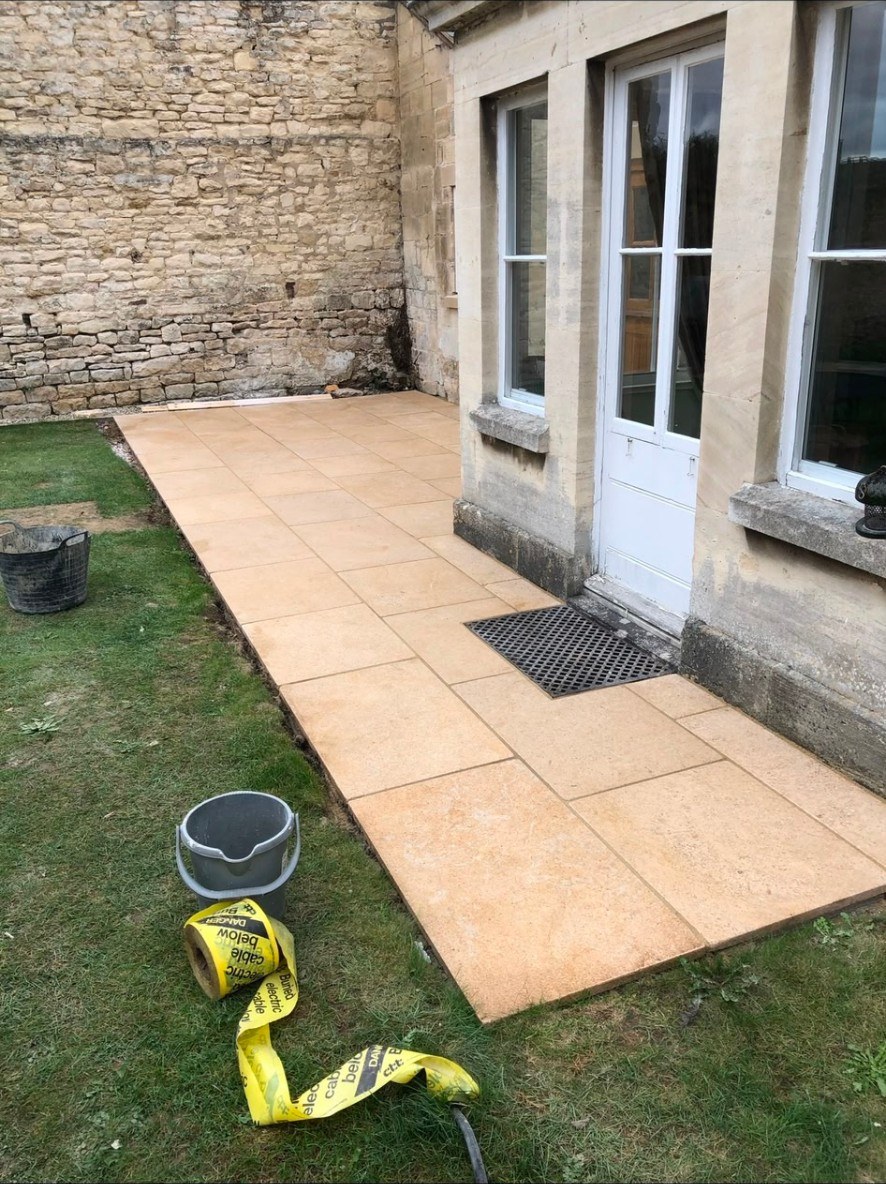 Newly laid buff natural stone paving slabs beside a Cotswold stone wall and period building.